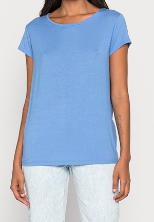 Frau trägt ein einfaches hellblaues T-Shirt mit kurzen Ärmeln und hellblaue Jeans, mit einem sichtbaren Blumen-Tattoo auf ihrem linken Unterarm.