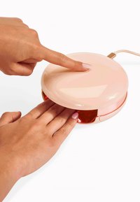 Różowe, błyszczące, okrągłe urządzenie z zaokrągloną pokrywką, wyświetlające czerwone światło wewnętrzne; ręka naciska na górną powierzchnię. Widać kabel.