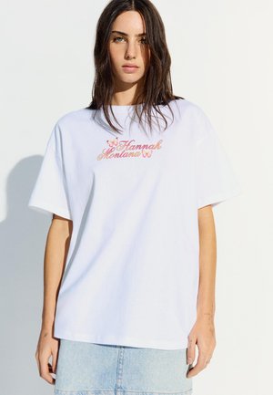 Jeune femme aux longs cheveux bruns portant un t-shirt blanc oversize avec le texte "Hannah Montana" et des motifs de papillons, jupe en denim clair.