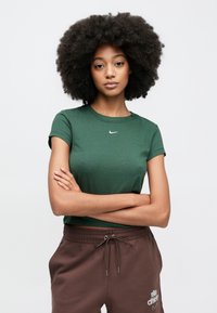 Πράσινο crop top μπλουζάκι από μαλακό βαμβάκι με κοντά μανίκια και μικρό λευκό λογότυπο Nike στο στήθος, συνδυασμένο με καφέ παντελόνι φόρμας με κορδόνι στη μέση.