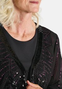 Schwarze durchsichtige Strickjacke mit einem filigranen Paillettenmuster in lila und schwarzen Tönen, mit einem V-Ausschnitt und einem glatten schwarzen Stoff darunter.