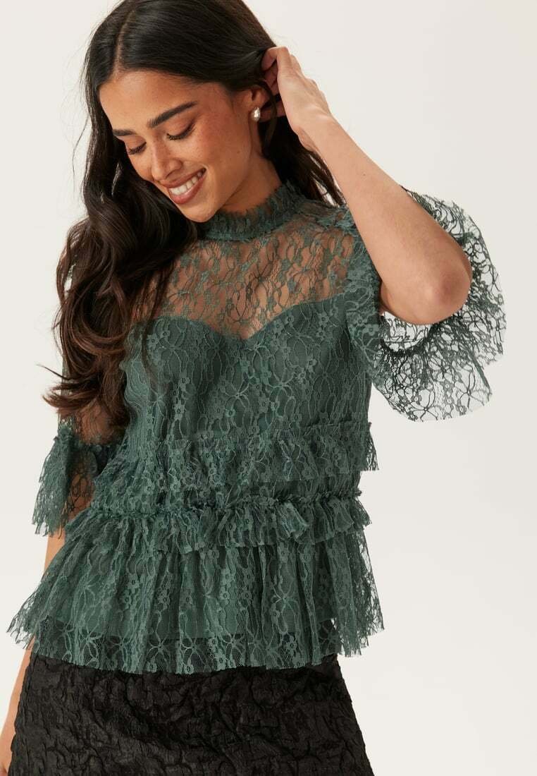 Top verde in pizzo con orlo e maniche arricciati. Tessuto trasparente con un intricato motivo floreale. Indossato su una gonna nera testurizzata.
