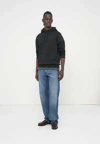 Zwarte katoenen hoodie met een kangaroezak, gedragen over een groen shirt, gecombineerd met losse blauwe jeans en donkere leren schoenen.