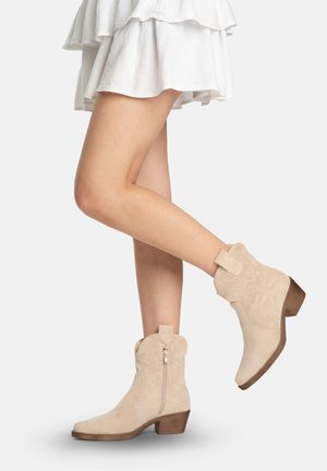 Ben med beige ruskinds-ankelstøvler med lave træsko og lynlåse i siden, kombineret med et hvidt lagdelt skørt.