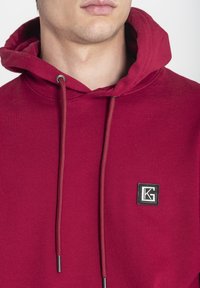 Sudadera de algodón roja con un bolsillo tipo canguro en el frente, capucha con cordón y un parche de logo cuadrado negro en el lado izquierdo del pecho. Textura suave.