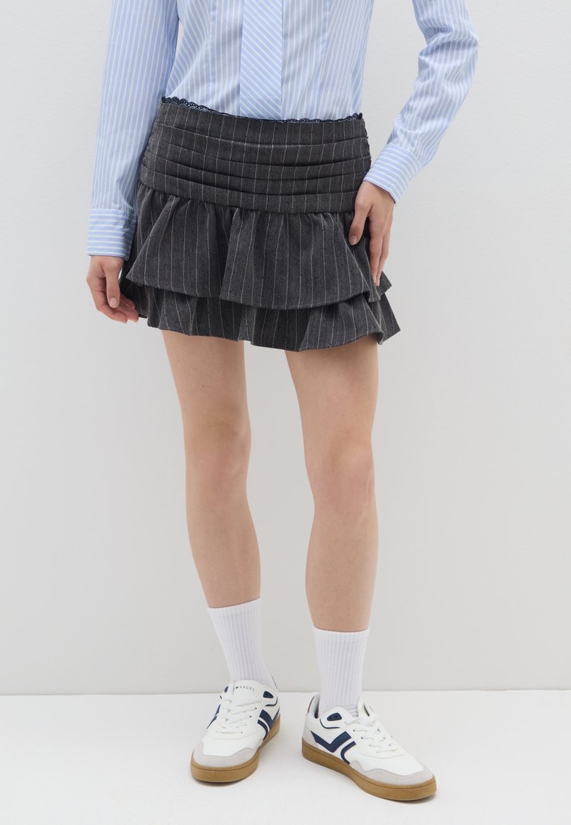 Zwarte minirok met zwarte streepjes en gelaagde ruches, met horizontale plooien bij de tailleband, gecombineerd met witte sneakers en kniekousen.