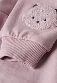 Rosa Stoffmanschette mit geripptem Bündchen und einem strukturierten Teddygesicht-Patch, das nahe dem Handgelenk aufgenäht ist.