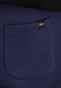 Gros plan sur une poche en tissu bleu marine avec une petite étiquette noire comportant un logo "A" stylisé en orange, cousue sur un fond de tissu plus foncé.