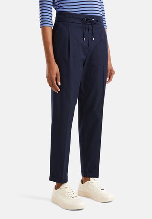 Kobieta w granatowych, wysokich spodniach z paskiem do wiązania, z podwiniętymi nogawkami, w białych trampkach i niebiesko-białym paskowanym topie.