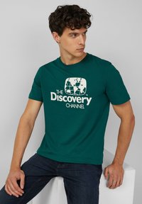 Szmaragdowa koszulka z krótkim rękawem wykonana z bawełny, z białym nadrukiem globu oraz napisem "The Discovery Channel" umieszczonym na środku z przodu.