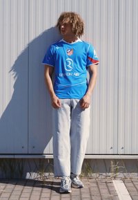 Blue athletic jersey with red accents and a logo, paired with light gray wide-leg pants and gray sneakers, standing against a textured backdrop.