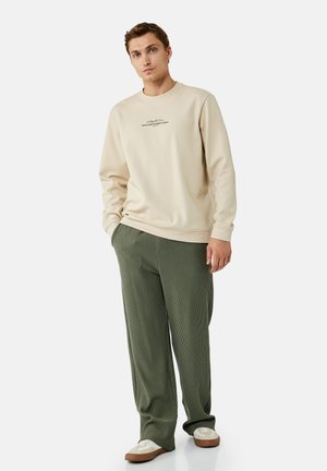 Ung mand iført en beige sweatshirt med tekst og løse grønne bukser står mod en ensfarvet lys baggrund.