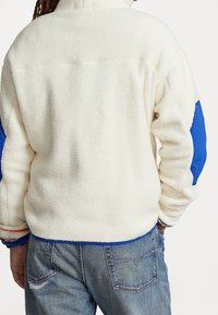 Vit fleecejacka med blå detaljer på ärmarna, med hög krage och texturerad tyg. Kombinerad med blå denimbyxor.