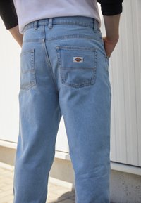 Light blue denim jeans with a relaxed fit, featuring two back pockets and a brand patch on the right pocket. Smooth texture.