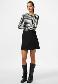 Femme les bras croisés, portant un haut à manches longues rayé noir et blanc, une jupe portefeuille noire et des bottes noires montant jusqu'aux genoux sur un fond blanc.