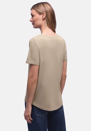 Kobieta o prostych blond włosach, ubrana w gładki beżowy T-shirt z krótkim rękawem i niebieskie dżinsy, pokazana od tyłu na białym tle.