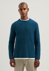 Blauwe textuurtrui met een ronde hals en lange mouwen, gedragen over een wit shirt, gecombineerd met lichtgekleurde broeken.