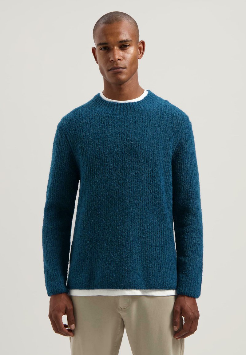 Blauwe textuurtrui met een ronde hals en lange mouwen, gedragen over een wit shirt, gecombineerd met lichtgekleurde broeken.