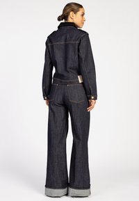Vrouw met een haar in een knot, gekleed in een donkerblauwe spijkerjack en wijde spijkerbroek, staand en met de rug naar de camera tegen een eenvoudige achtergrond.