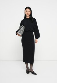 Fekete oversize pulóver, testhezálló fekete midi szoknya, fekete áttetsző harisnya és hegyes orrú fekete magassarkú. Kígyóbőr mintás táska az ölében.