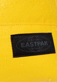 Material impermeable amarillo con una superficie texturizada, que presenta una etiqueta de tela negra con el logo "EASTPAK" y un diseño de globo.