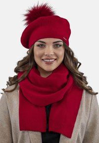 Berretto a maglia rosso con un pom-pom soffice e sciarpa rossa coordinata, entrambi caratterizzati da una texture morbida e un design semplice, abbinati a un cappotto beige.