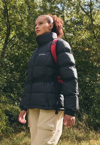 Czarna kurtka puchowa z wysokim kołnierzem, regulowanym paskiem i logo. Połączona z beżowymi spodniami cargo z boczną kieszenią. Widoczny pasek czerwonego plecaka.