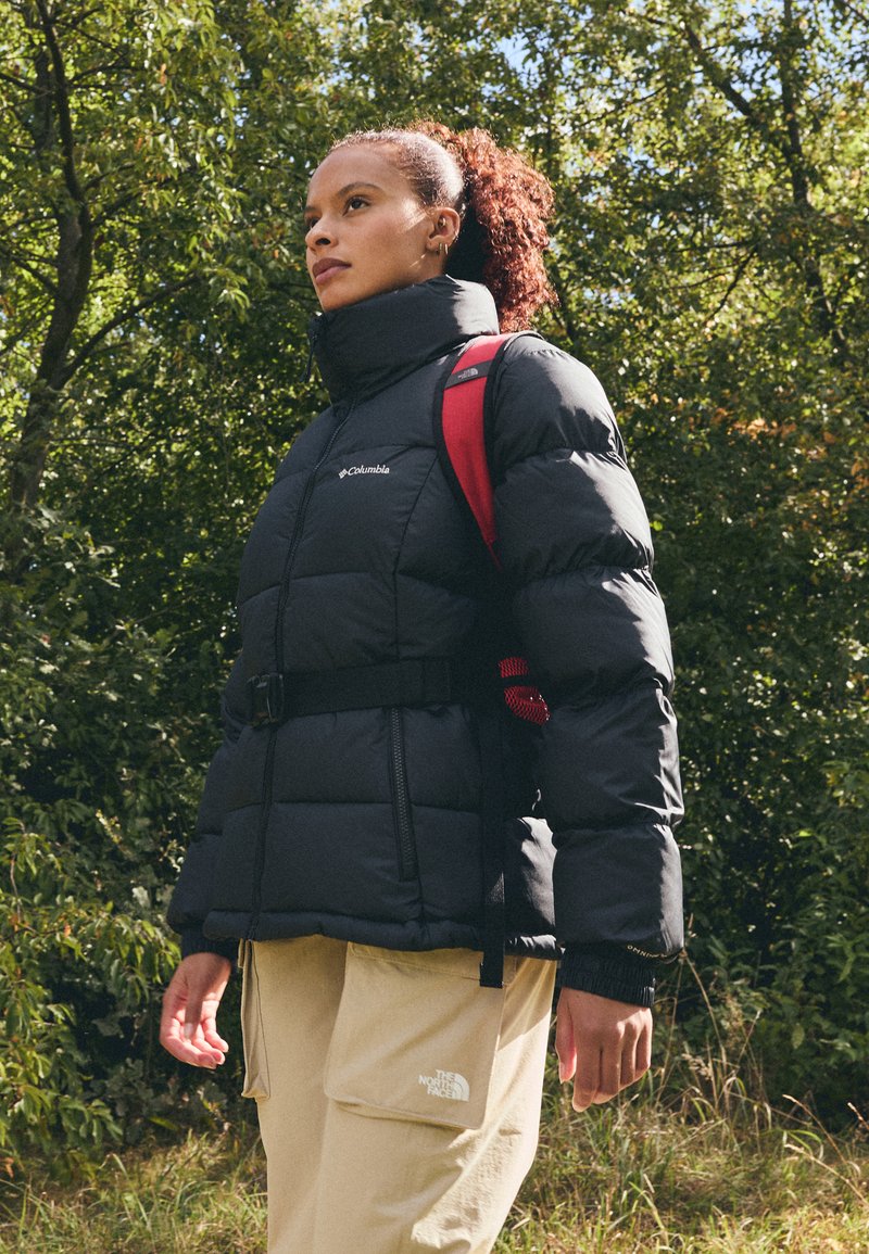 Czarna kurtka puchowa z wysokim kołnierzem, regulowanym paskiem i logo. Połączona z beżowymi spodniami cargo z boczną kieszenią. Widoczny pasek czerwonego plecaka.