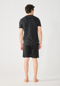 Black, short-sleeve t-shirt with a textured pattern, paired with plain black shorts. Model stands barefoot, facing away from the camera.