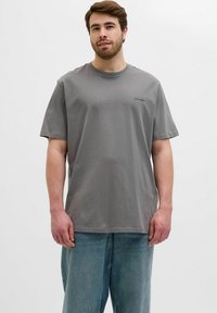 Camiseta de algodón gris con mangas cortas y cuello redondo. Presenta un pequeño parche con logo en el pecho. Se lleva con unos jeans sueltos de color azul claro.