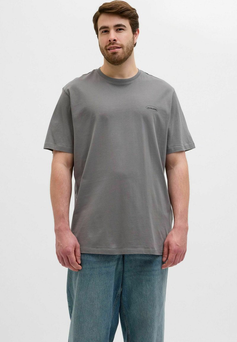 Camiseta de algodón gris con mangas cortas y cuello redondo. Presenta un pequeño parche con logo en el pecho. Se lleva con unos jeans sueltos de color azul claro.