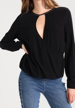 Kvinde iført en sort langærmet top med en frontnøgleudskæring og blå jeans med snøredetalje i siden.