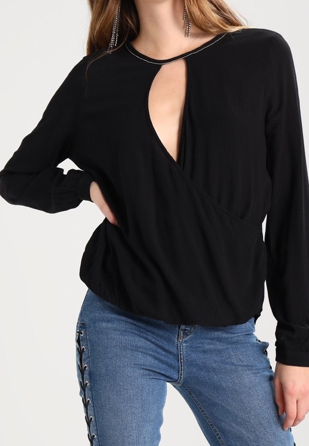 Femme portant un haut noir à manches longues avec une découpe en forme de goutte à l'avant et un jean bleu avec un détail à lacets sur le côté.