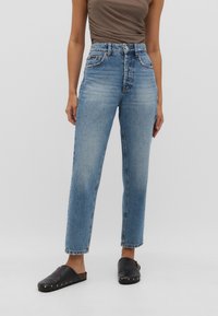 Mujer vestida con jeans azules de tiro alto y pierna recta, combinados con zapatos negros sin cordones y una blusa sin mangas marrón.