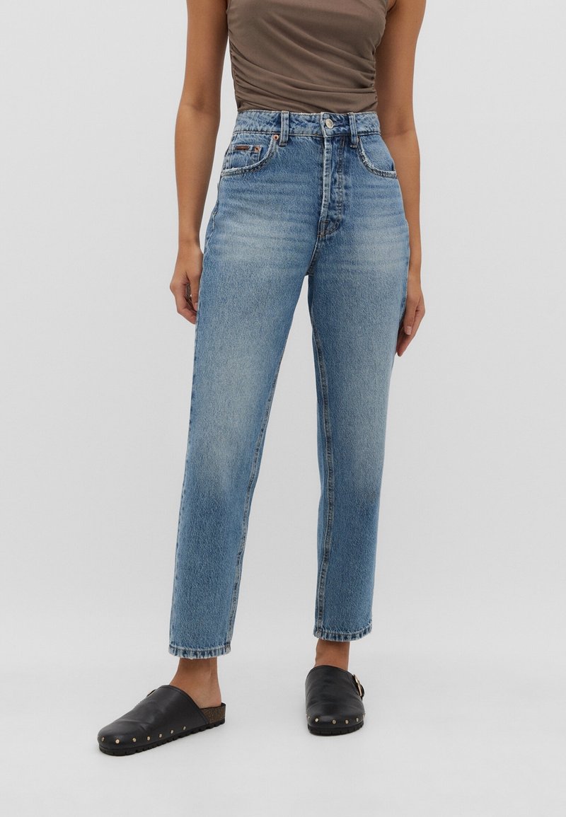 Mujer vestida con jeans azules de tiro alto y pierna recta, combinados con zapatos negros sin cordones y una blusa sin mangas marrón.