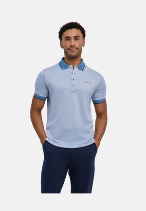 Mann trägt hellblaues Poloshirt mit dunkelblauem Kragen und Ärmelsaum, steht mit den Händen in den Taschen der dunkelblauen Hose.
