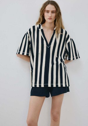 Kobieta stoi z rękami za plecami, ubrana w oversize czarno-białą, pionowo paskowaną koszulę z krótkim rękawem i czarne szorty na jednolitym tle.