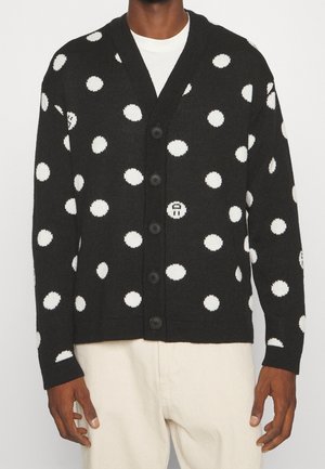 Cárdigan negro abotonado con grandes lunares blancos usado sobre una camisa blanca y combinado con pantalones de color claro.