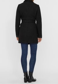 Schwarzer, mittellanger Mantel aus Wildleder mit tailliertem Schnitt und langen Ärmeln, kombiniert mit dunkelblauen Skinny-Jeans und schwarzen Knöchelstiefeln.