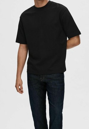 Homme portant un t-shirt noir à manches courtes et un jean bleu foncé, debout devant un fond clair uni.