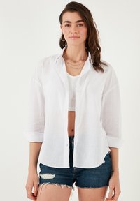 White button-up shirt, lightweight linen fabric, long sleeves rolled up, paired with a fitted white tank and denim shorts. Subtle texture visible.