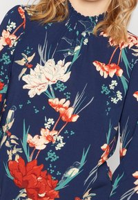 Blus med blommönster på marinblå bakgrund, prydd med röda, vita och gröna blommor, volangkrage och långa ärmar.