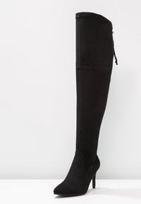 Černé z mikroplyše vysoké kozačky na jehlovém podpatku s špičatou špičkou a detailem krajkového zavazování na zadní straně, postavené na bílé ploše proti jednoduchému pozadí.