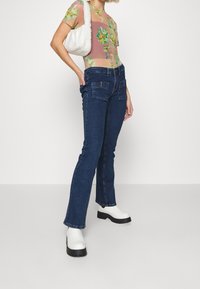 Kvinna som bär en blommig transparent topp, mörkblå jeans med framfickor, vita kraftiga stövlar och bär en vit fluffig handväska över axeln.