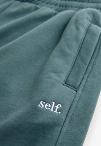 Prim-plan cu buzunarul pantalonilor de trening verde-albăstru cu talie elastică și text alb brodat „self.” pe o țesătură moale.