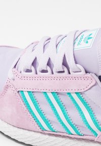 Chaussure de sport lilas clair avec un revêtement en daim texturé, trois bandes aqua et des lacets plats ; tige en synthétique lisse et semelle texturée.