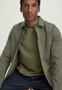 Olivegrüner, leichter Blouson mit sanfter Oberfläche, Knopfverschluss und langen Ärmeln über einem passenden Rundhalspullover. Darunter sind dunkle Hosen zu sehen.