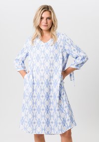 Helles blaues knielanges Kleid mit einem auffälligen weißen geometrischen Muster, V-Ausschnitt, dreiviertellangen Ärmeln und seitlichen Taschen. Glatte Stoffoberfläche.