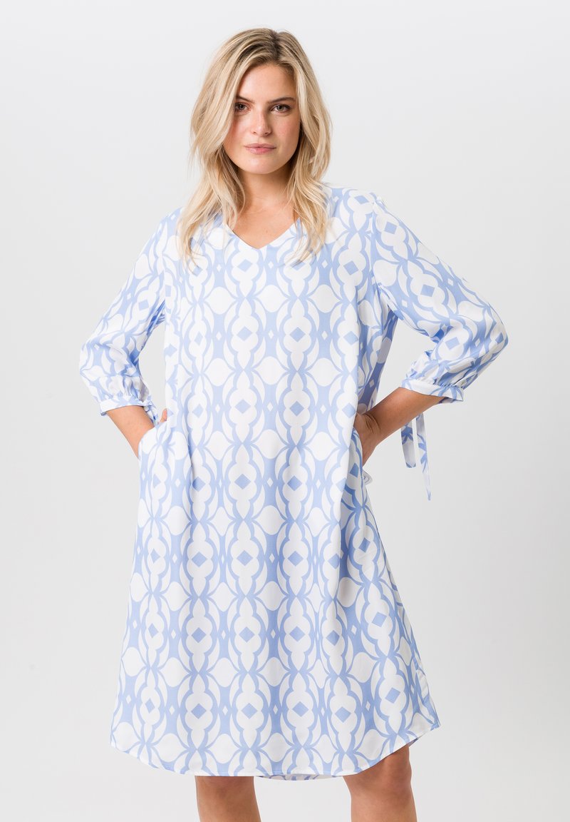 Helles blaues knielanges Kleid mit einem auffälligen weißen geometrischen Muster, V-Ausschnitt, dreiviertellangen Ärmeln und seitlichen Taschen. Glatte Stoffoberfläche.