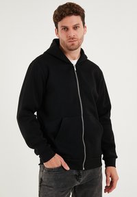 Černá mikina na zip s přední kapsou, měkkou látkou, nastavitelnou kapucí se stahovací šňůrkou a žebrovanými manžetami, nošená přes bílé tričko.